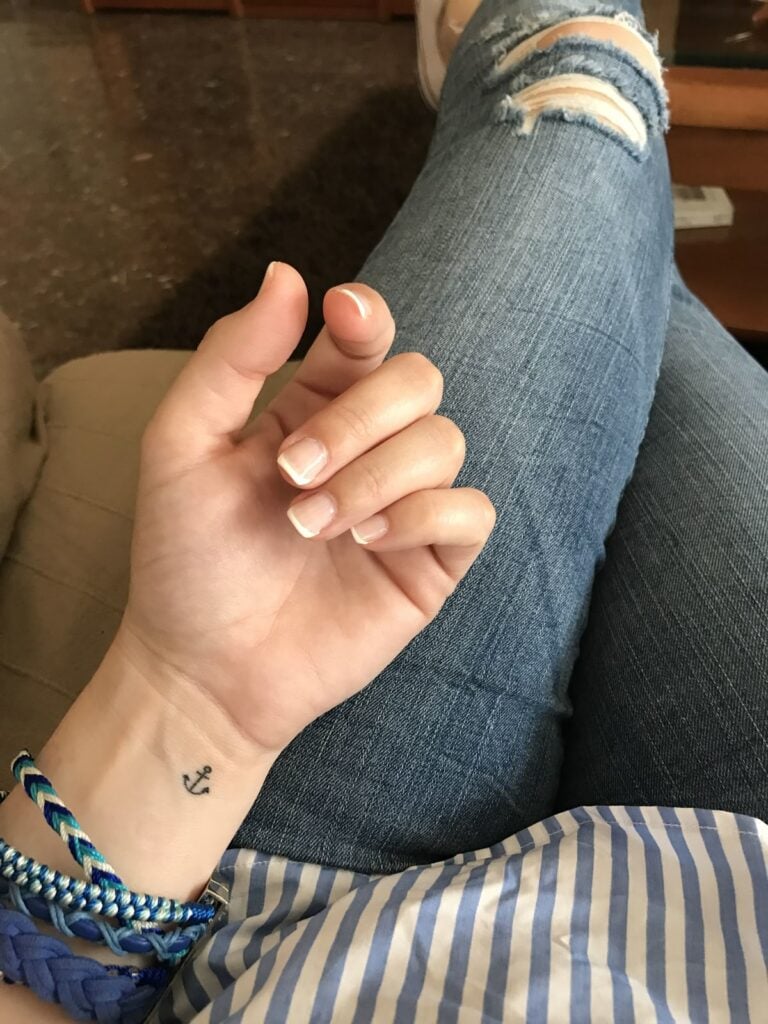 Close-up of hand with French manicure, small anchor wrist tattoo and blue braided bracelets resting on ripped blue jeans and striped shirt.