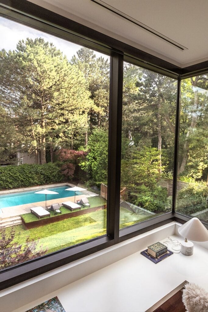 Interior desk view through large glass window to backyard pool with loungers, umbrellas and lush wooded garden — modern home oasis.