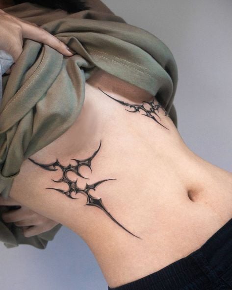 Side torso close-up showing black abstract tribal-style ribcage tattoos beneath a lifted green shirt.