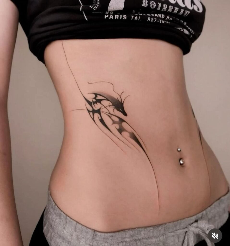 Abstract black tribal tattoo on side torso and slim waist with belly button piercing, cropped top and low-rise pants