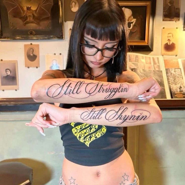 Woman with glasses showing script forearm tattoos 'Still Strugglin' and 'Still Shyin' in a tattoo studio, script forearm tattoo inspiration