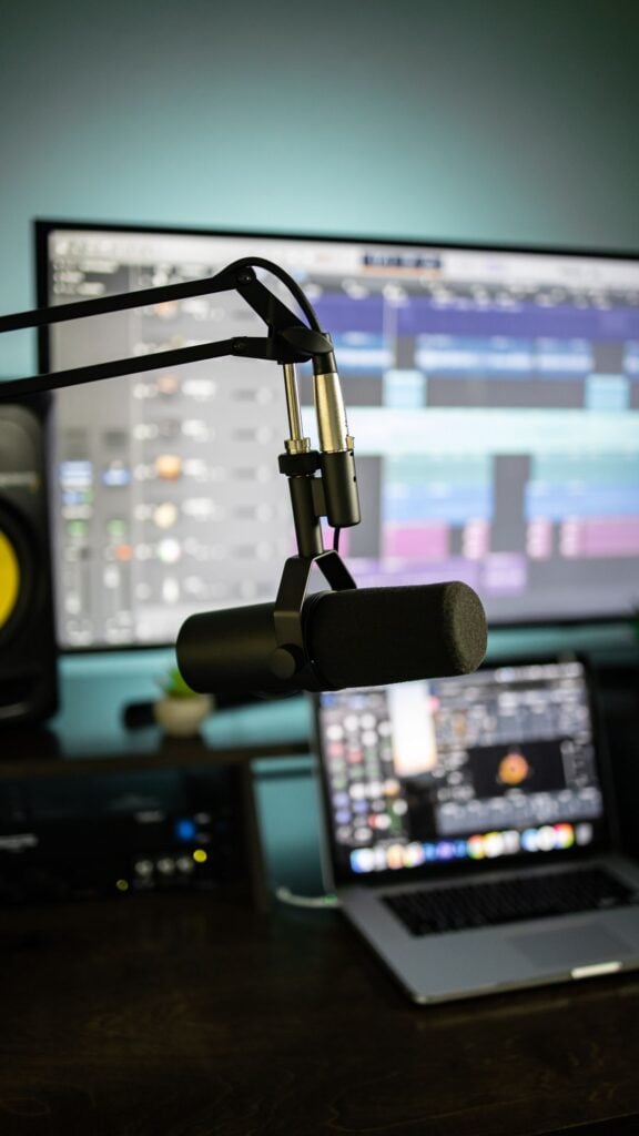 Podcast microphone on adjustable boom arm before laptop and monitor displaying audio-editing software, home studio setup