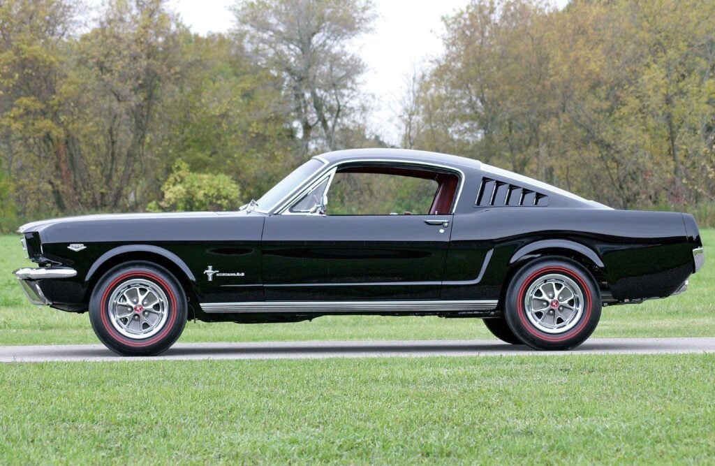 Black classic Ford Mustang fastback side profile with redline wheels parked on roadside grass, vintage American muscle car