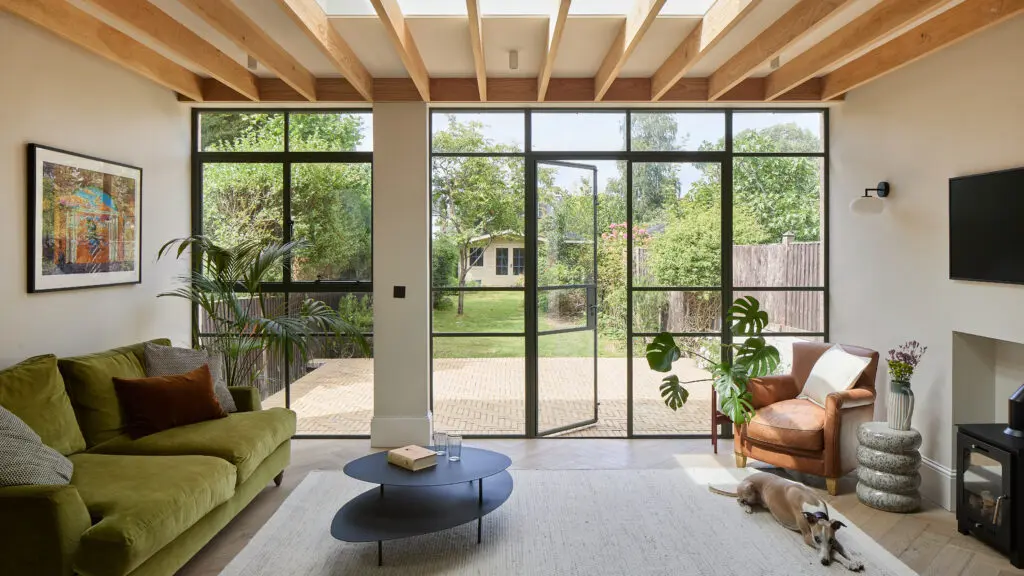 Modern living room with floor-to-ceiling black-framed glass doors to garden, green velvet sofa, leather armchair, dog