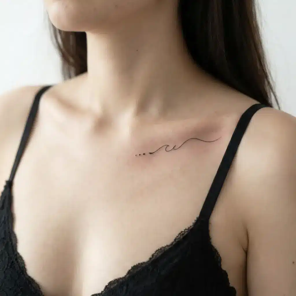 Minimal fine-line wave tattoo on woman's collarbone above black lace bralette, delicate chest ink with shoulder strap detail