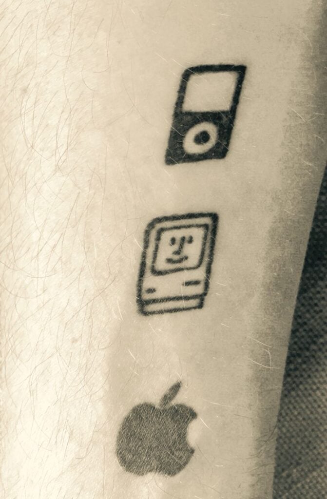Close-up forearm tattoo of three black retro Apple icons: iPod, smiling Macintosh face, and bitten Apple logo on light skin.