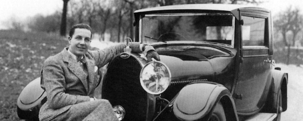 Black-and-white photo of a man in a suit leaning on a vintage luxury car from the 1920s, classic automobile on a tree-lined road