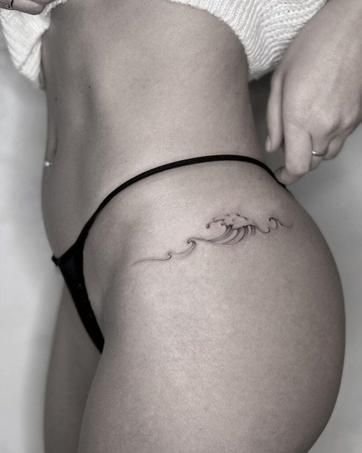 Close-up of a minimalist wave tattoo on a person's hip/upper thigh, wearing a black thong and cream sweater; delicate fine-line design.