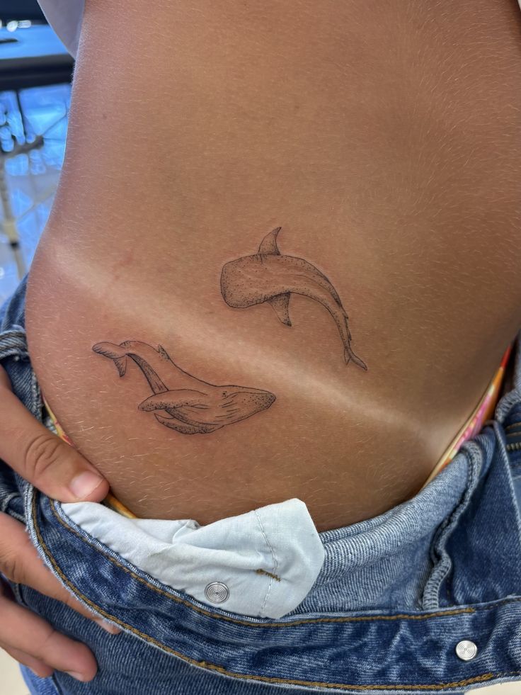 Minimal fine-line twin whale tattoo on hip above jeans — small ocean-themed lower abdomen ink