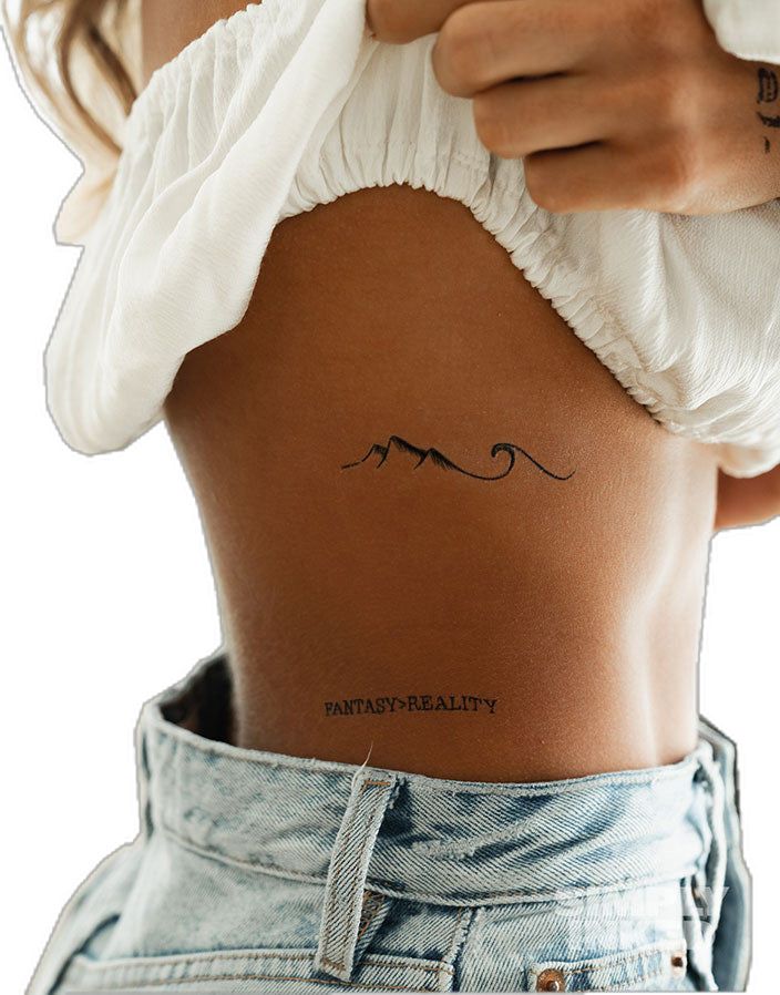 Minimal mountain-and-wave rib tattoo above 'FANTASY-REALITY' text on model's side, white crop top and high-waisted denim jeans