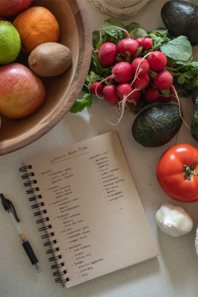 Weekly meal plan notebook and grocery list surrounded by fresh produce: radishes, avocados, tomato, garlic and bowl of apples, kiwi, oranges.