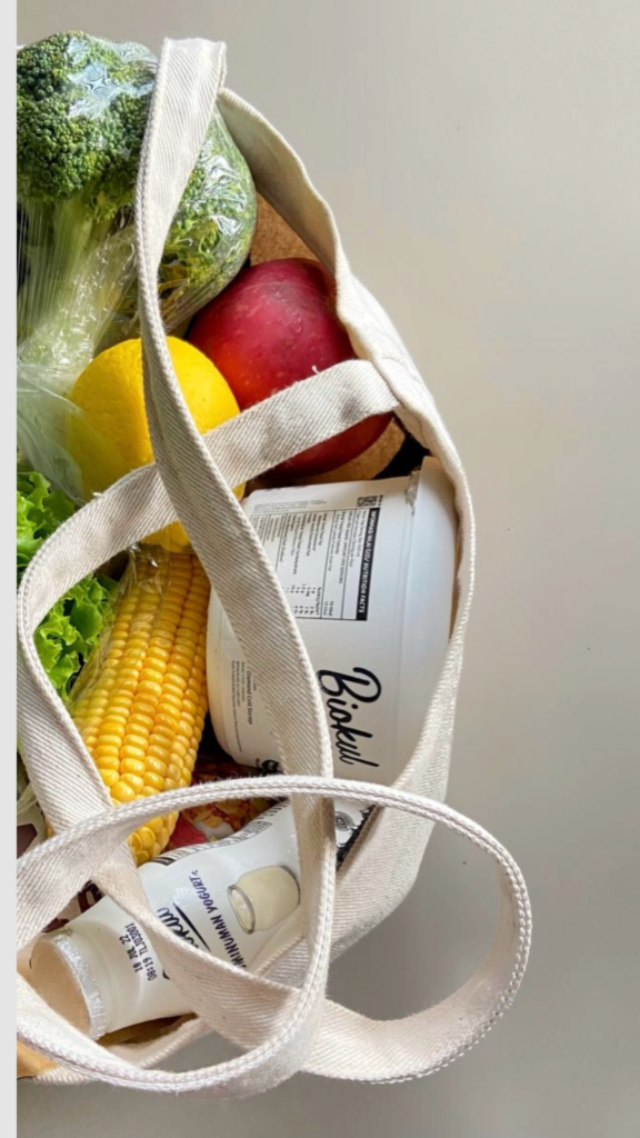 Reusable canvas grocery bag filled with fresh produce: broccoli, corn, apple, lemon, lettuce and yogurt