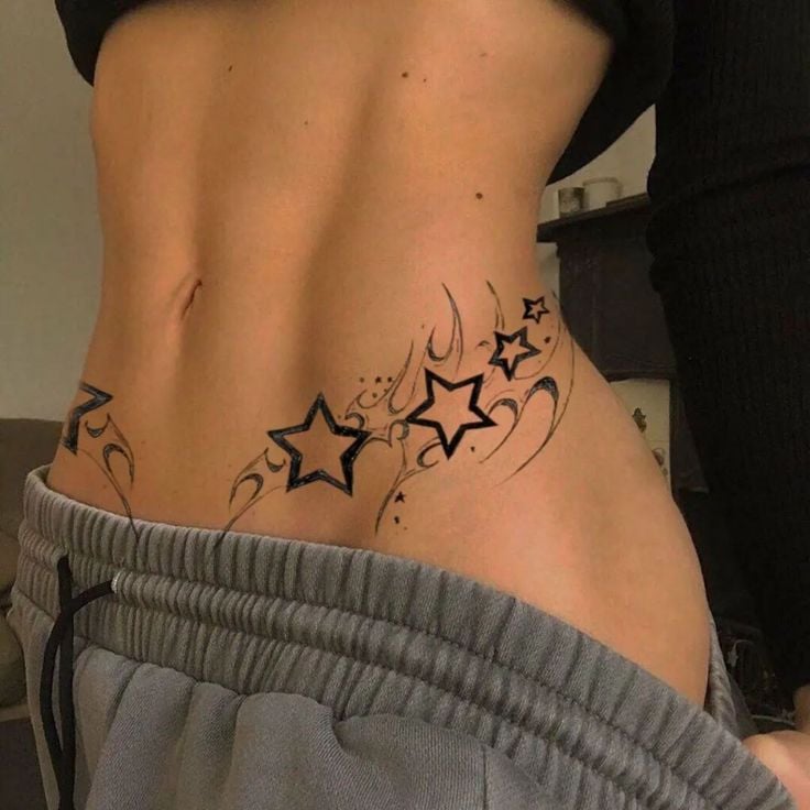 Close-up of lower torso with black star and tribal-style hip tattoo across abdomen, exposed midriff and gray sweatpants waistband.