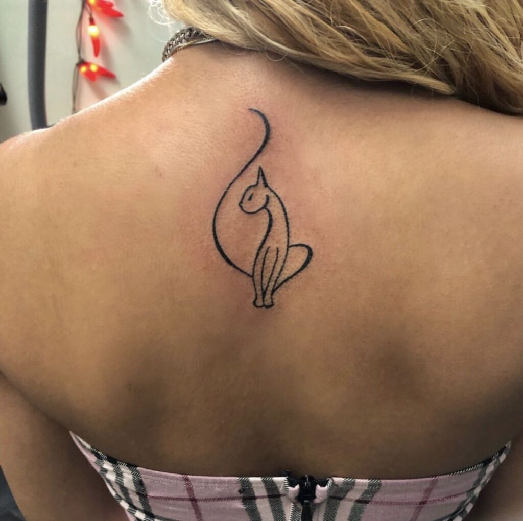 Small minimalist fine-line black cat tattoo centered on upper back between shoulder blades on person wearing a strap top
