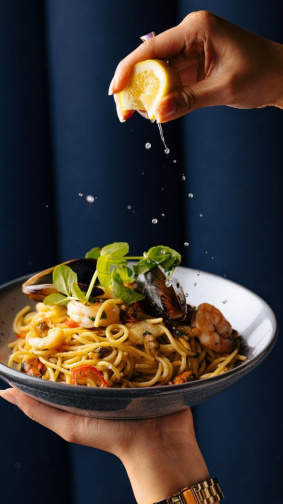 Seafood spaghetti with shrimp and mussels in a bowl, hand squeezing lemon over pasta with fresh herbs and bright citrus drizzle