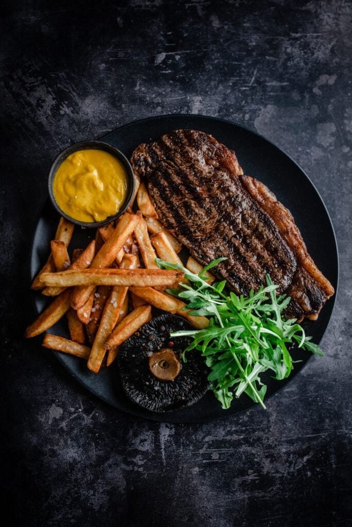 Grilled ribeye steak with golden fries, roasted portobello, arugula and mustard dipping sauce on black plate