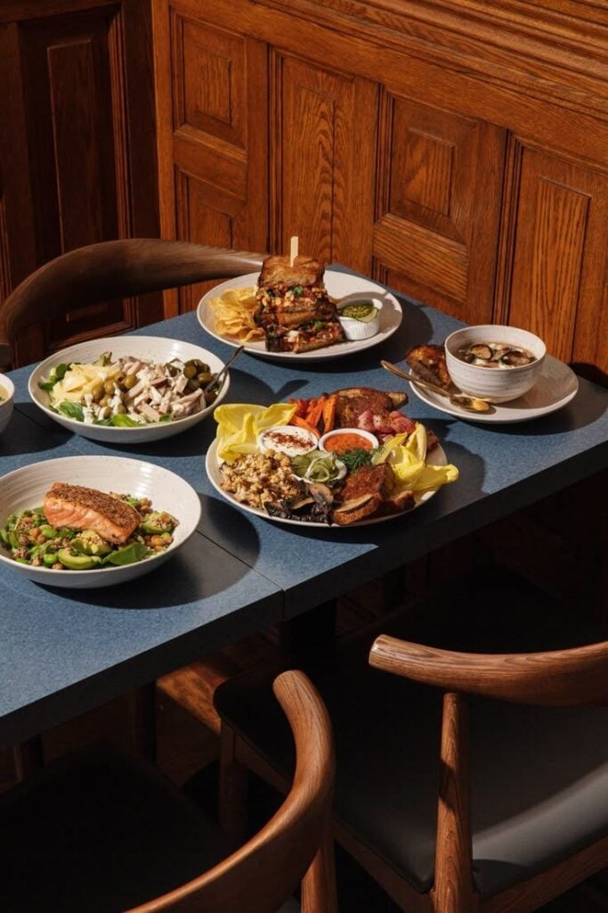 Gourmet brunch spread on blue table: grilled salmon salad, stacked sandwich, mixed salads, soup and roasted vegetables in wood-paneled dining room