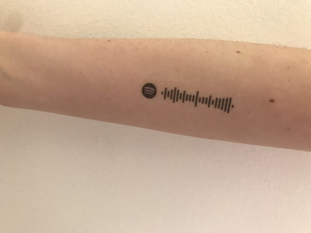 Close-up forearm tattoo of a Spotify scannable code and logo on light skin against a white background