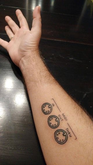 Forearm tattoo of three MIDI DIN ports labeled IN, OUT, THRU - music/electronics themed on dark wood background