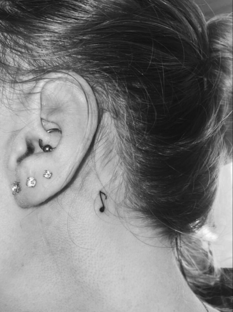 Black-and-white close-up of ear with three stud piercings, hoop cartilage piercing and small music-note tattoo behind ear