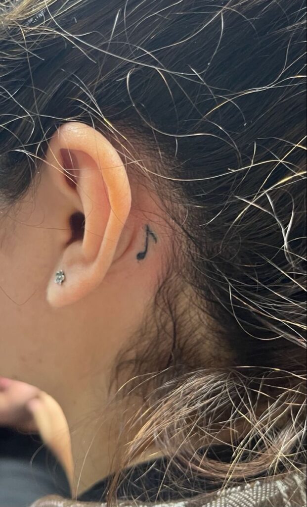 Close-up of ear with small black music-note tattoo behind ear and silver stud earring, dark hair strands