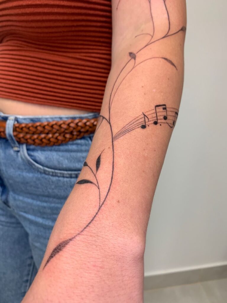 Delicate black-ink forearm tattoo: swirling vine with leaves and musical notes on a staff