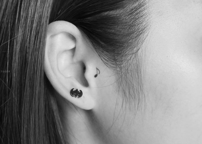 Black-and-white close-up of ear with small music-note tattoo behind ear and black bat-shaped stud earring.