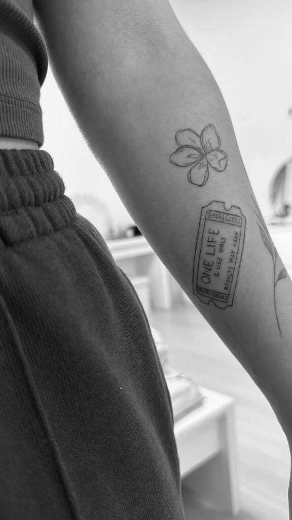 Black and white close-up of forearm tattoo: minimalist flower and One Life ticket design