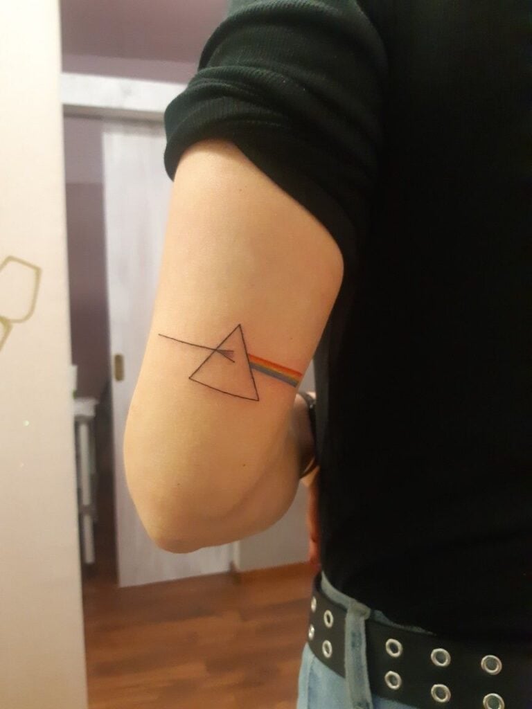 Minimal prism tattoo with rainbow spectrum (Pink Floyd-inspired) on inner upper arm of person wearing black shirt