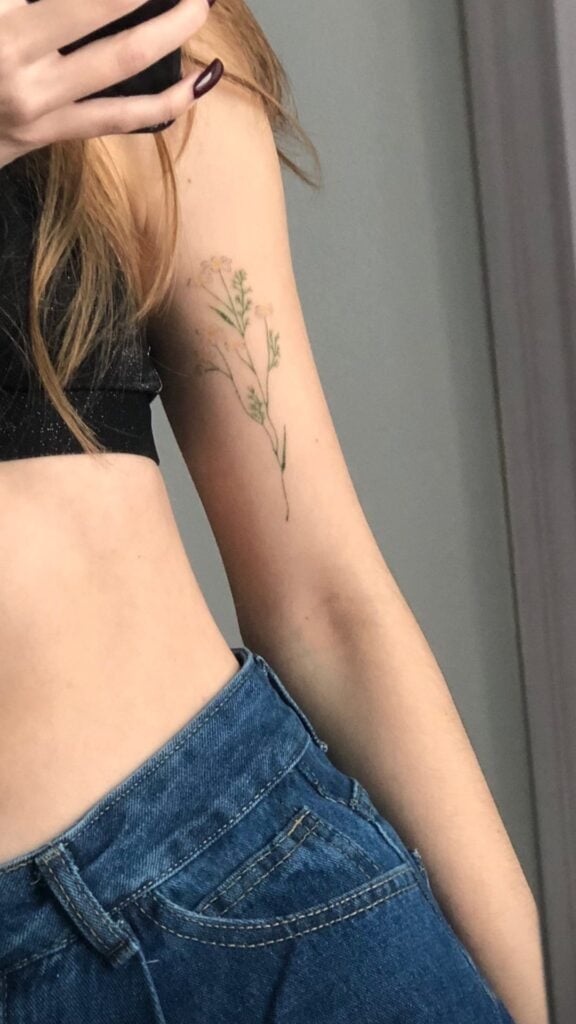 Wildflower arm tattoo mirror selfie: person in black crop top and blue high-waisted jeans showing delicate botanical ink on inner upper arm.