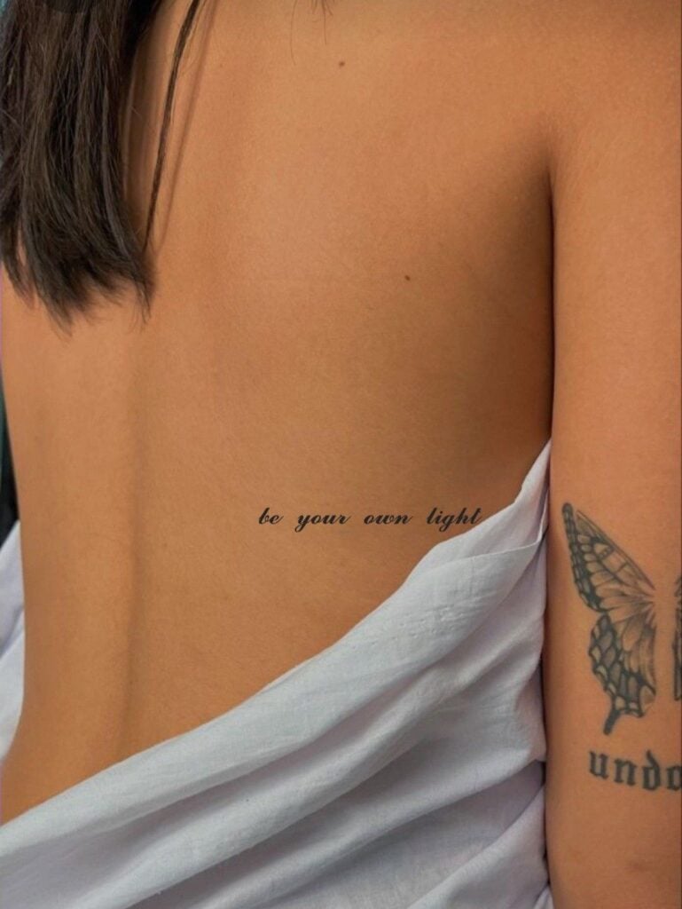 Close-up of person's side with small script tattoo 'be your own light', butterfly arm tattoo, draped white sheet