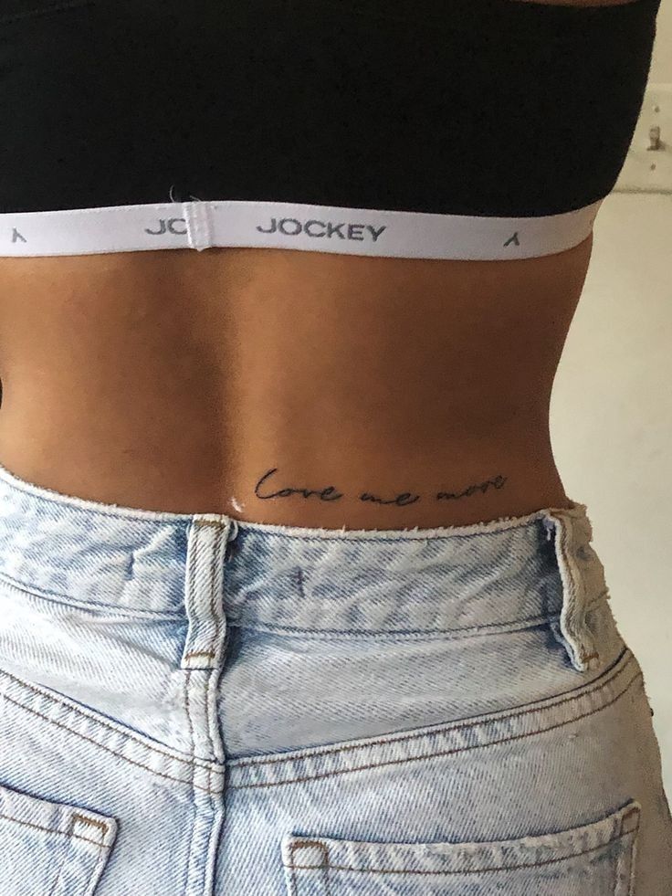Close-up of woman's lower back with cursive 'love me more' tattoo above high-waisted light denim jeans and bralette