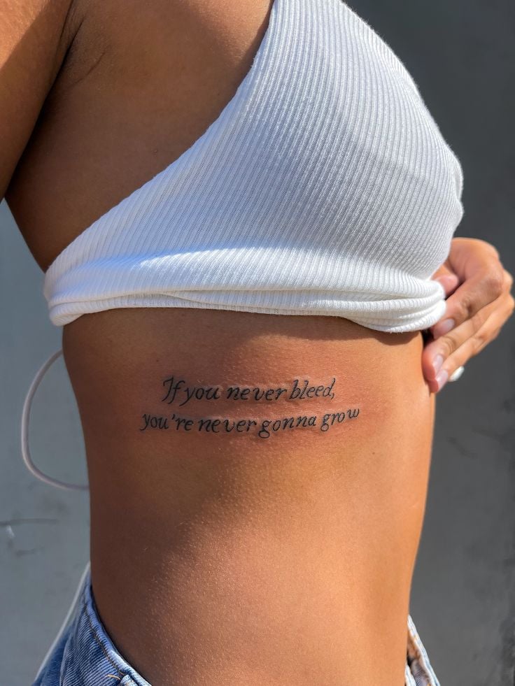 Rib-side script tattoo reading If you never bleed, you're never gonna grow on woman wearing white crop top