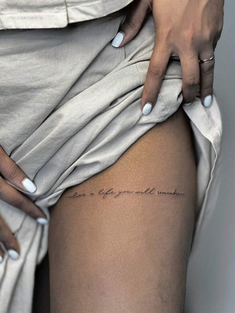 Minimalist thigh script tattoo reading live a life you will remember with white-manicured nails and beige fabric