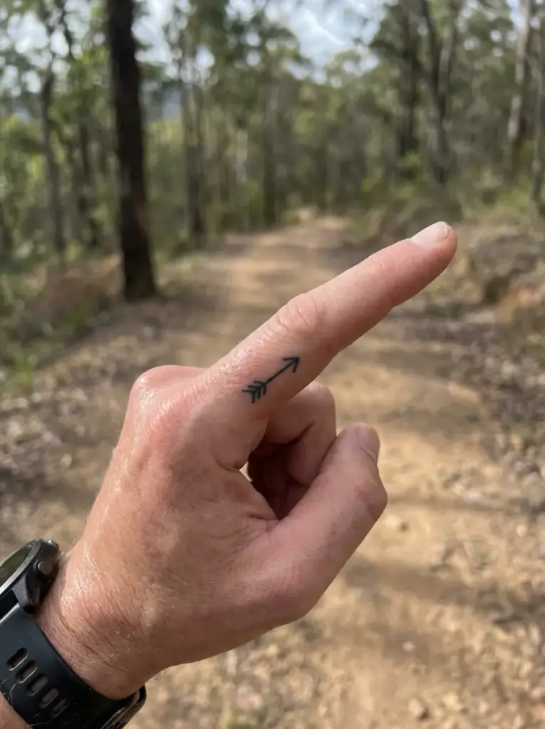 Arrow finger tattoo on hand pointing along a sunlit dirt hiking trail through a forest