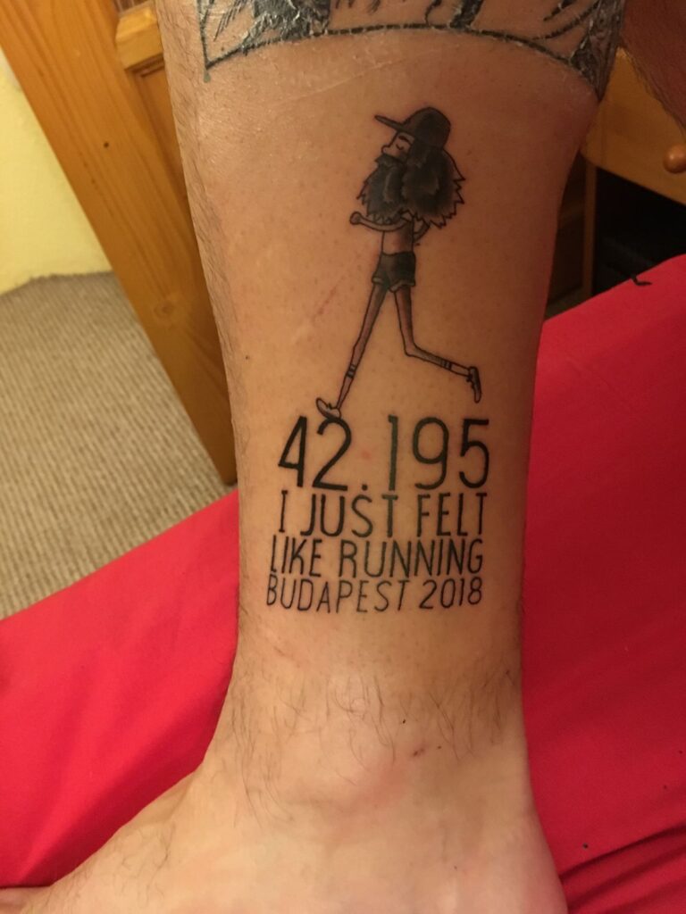 Ankle tattoo of a bearded cartoon runner and text '42.195 I just felt like running Budapest 2018' commemorating a marathon.