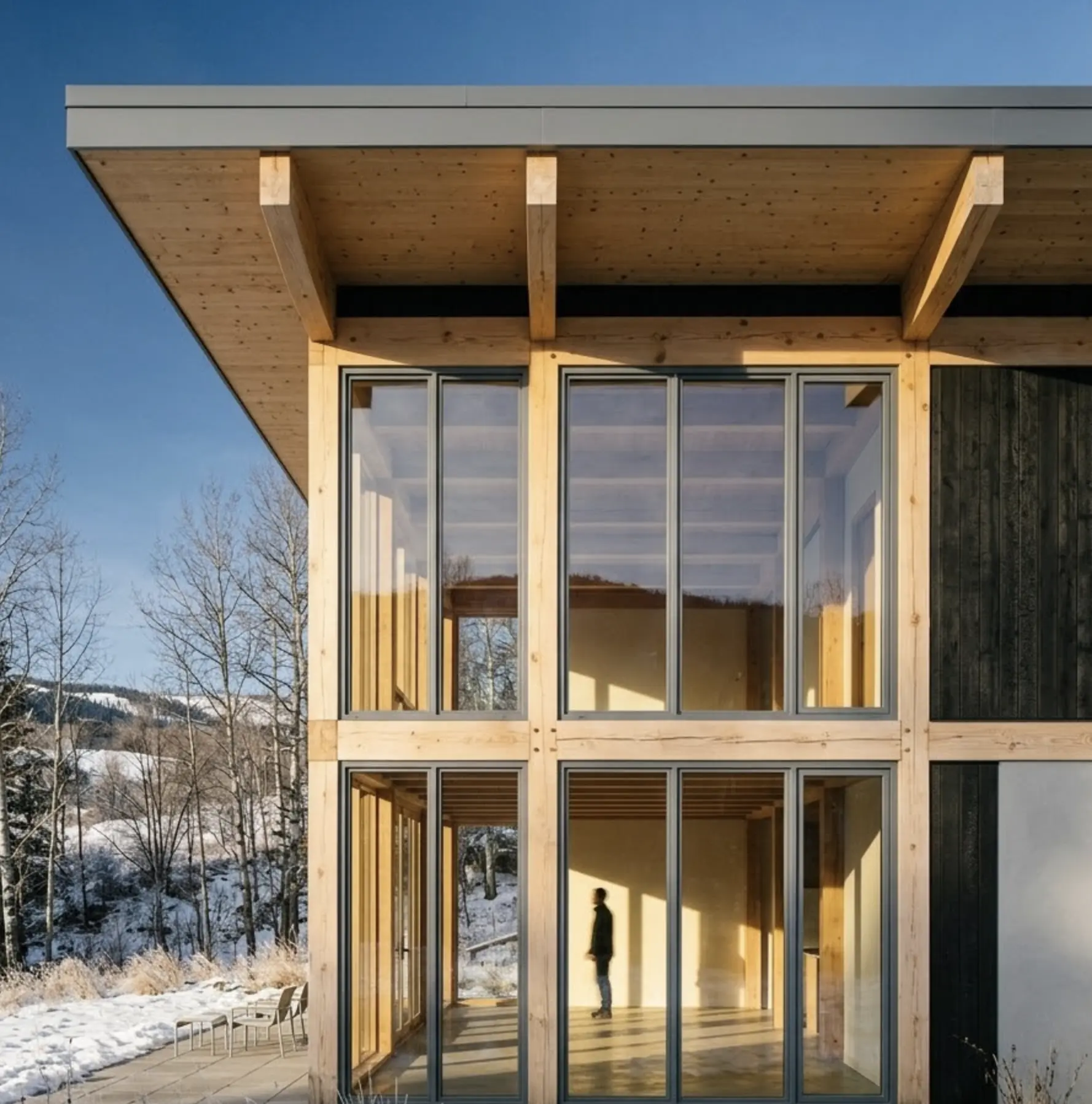 Modern timber-frame home with large glass facade and floor-to-ceiling windows overlooking snowy landscape, person silhouette