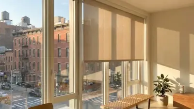 Sunlit modern apartment with large windows, roller shades, bench and potted plant overlooking urban street