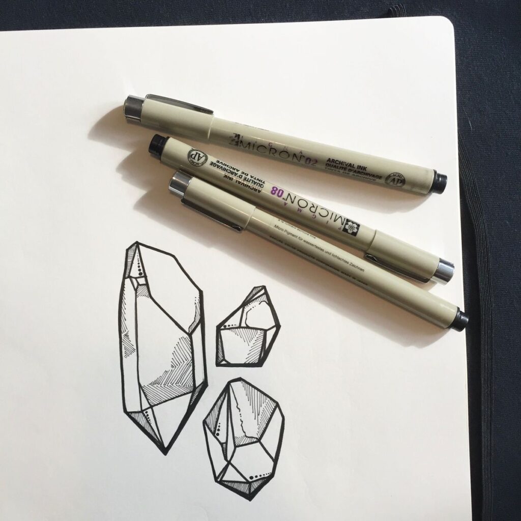 Sakura Pigma Micron pens on a sketchbook beside black ink geometric crystal sketches