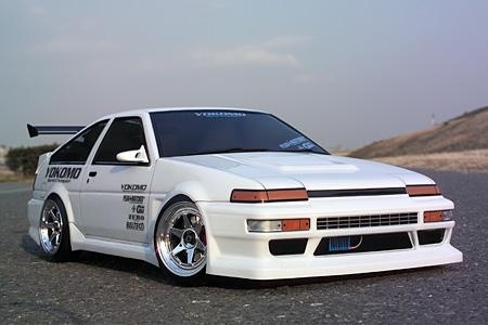 White Toyota AE86 Trueno drift car with wide body kit, chrome wheels and rear wing parked on asphalt road