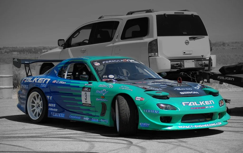Teal Falken-sponsored Mazda RX-7 drift car with large rear wing and sponsor decals, parked at a racetrack