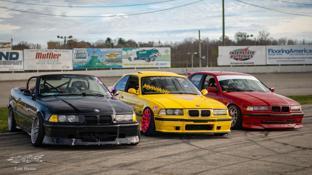 Three modified BMW drift cars (black, yellow with pink wheels, red) lined up on a racetrack for a drift event