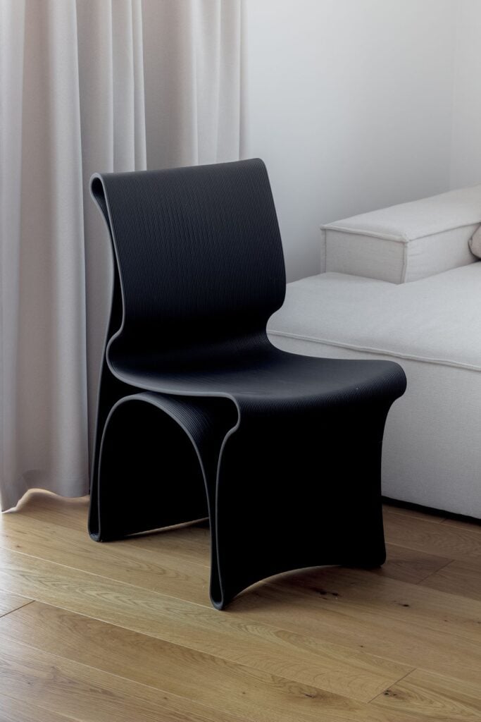 Modern black sculptural chair next to light sofa in minimalist living room with hardwood floor and neutral curtains