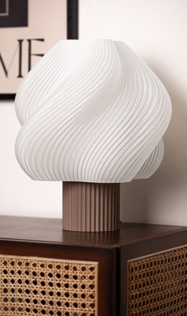 Modern white sculptural swirl table lamp with ribbed brown base on wooden rattan cabinet, minimalist decor.