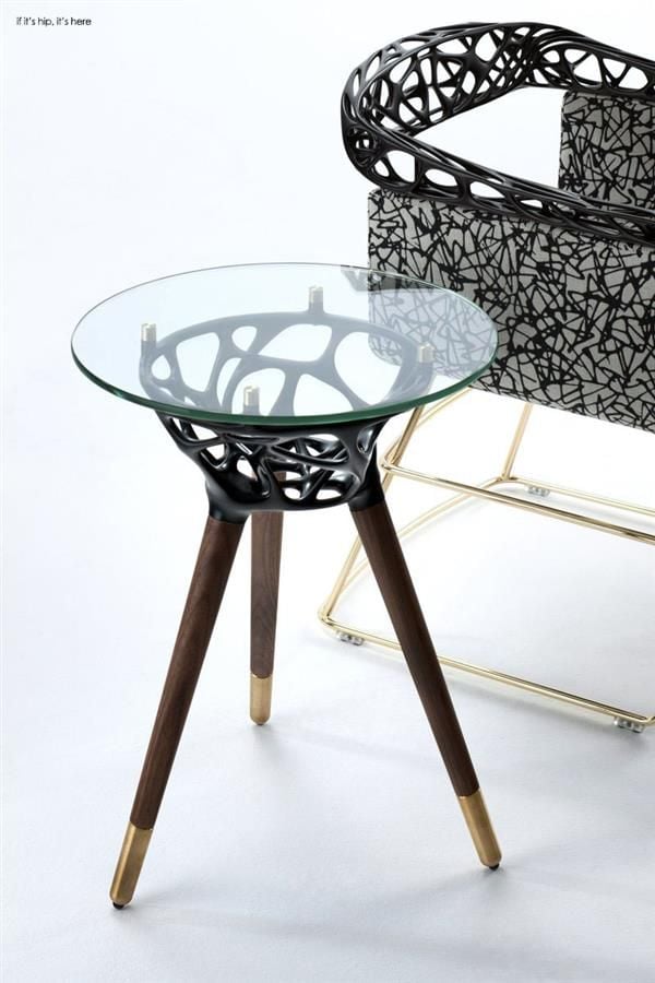 Modern glass-top side table with sculptural black lattice base, walnut legs and brass caps, staged beside patterned chair.