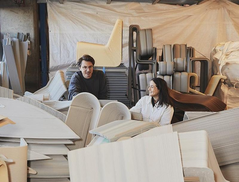Two furniture designers examining stacked molded-plywood chair prototypes in a busy workshop studio
