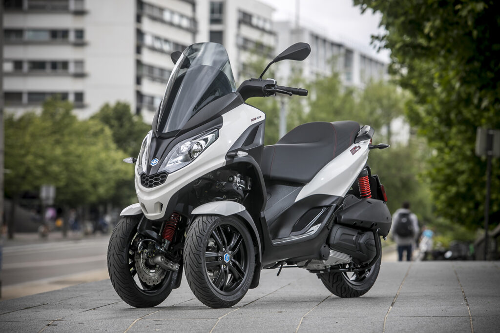Three-wheeled white Piaggio MP3 scooter parked on city sidewalk, front view with buildings and trees behind