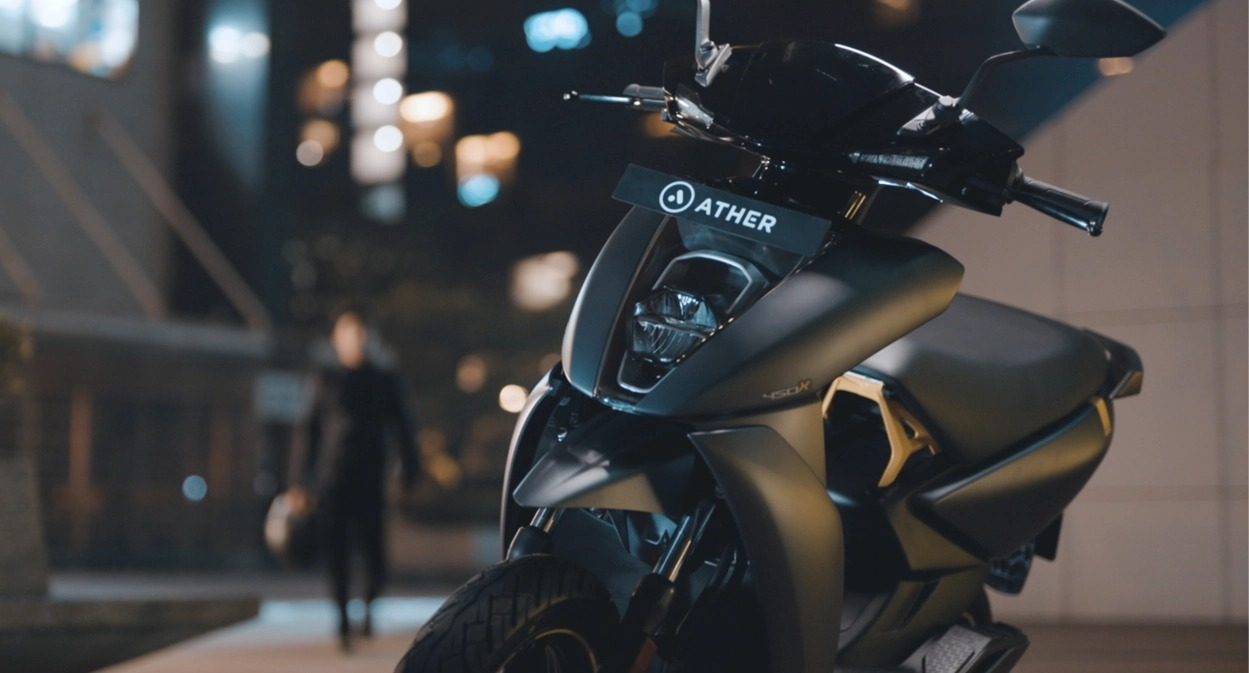 Close-up of matte black Ather electric scooter parked in urban night setting with blurred pedestrian and city lights