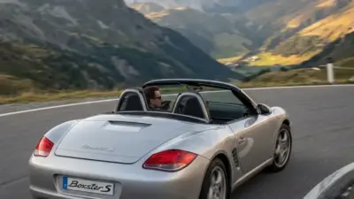 Silver Porsche Boxster S convertible driving on winding mountain road with scenic alpine valley backdrop