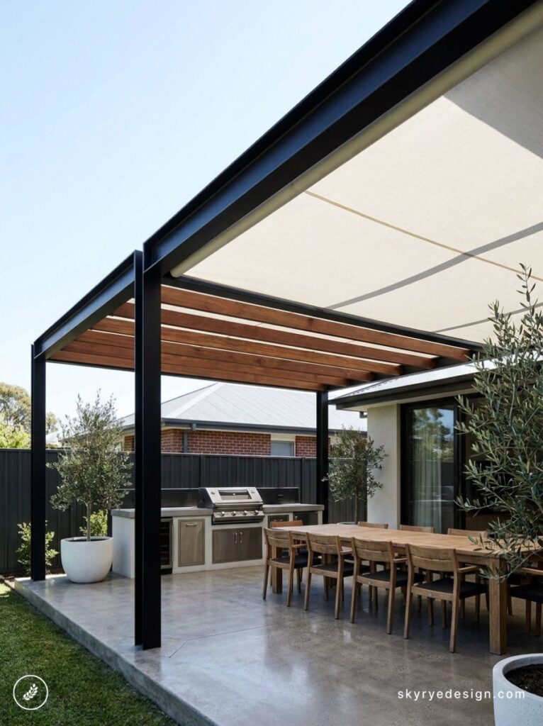 Outdoor backyard patio under a modern pergola with a long wooden dining table, chairs, and a built-in barbecue grill.